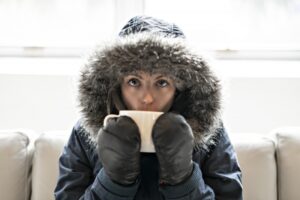 cold-person-indoors-wearing-a-hooded-coat-and-mittens-and-drinking-hot-tea